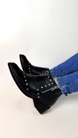 Botín corto de cuero negro con textura combinada en la puntera y el talón. Cuenta con punta fina, apliques metálicos en forma de tachas y cierre lateral plateado.