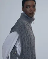 Chaleco largo de punto trenzado color gris con cuello alto y cierre frontal con cremallera metálica.