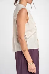 Blusa sin mangas de color beige, confeccionada en tela ligera con detalles de bordados florales frontales, encajes y cierre delantero con tiras para anudar.