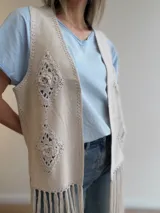 Chaleco de gamuza sintética color marrón, sin mangas, con diseño abierto, bordados geométricos de crochet en los laterales y largos flecos en el ruedo.