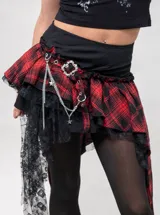 Minifalda asimétrica de cuadros tartán en rojo y negro, con detalles de encaje negro, una cadena metálica colgante y hebilla decorativa.