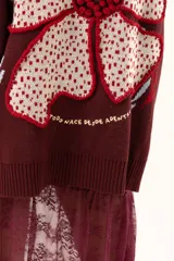 Saco de punto color vino con diseño holgado, cuello redondo y mangas largas. Presenta un gran bordado central en la espalda con forma de flor en tonos crema y rojo, acompañado de un pequeño bordado de sol y una frase bordada en la parte inferior.