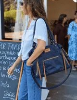 Cartera tipo bolso de cuero azul marino, con detalles y tiras de cierres en color tostado. Cuenta con bolsillos externos con cierre, asas dobles para hombro y correa larga ajustable.