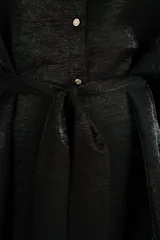Camisa larga de satén negro, fluida, con cuello camisero, cierre frontal de botones dorados, mangas tres cuartos y lazo ajustable en la cintura.