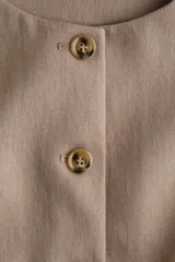 Chaleco color tostado con cuello redondo, sin mangas y cierre frontal con botones oscuros. Presenta pinzas frontales para un calce estructurado y terminación inferior en picos divididos.