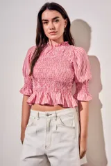 Blusa de algodón con diseño a rayas verticales en tonos rosa y blanco. Presenta un cuerpo con fruncido elástico (smock), cuello alto con volados y mangas tres cuartos abullonadas con terminación en volado.