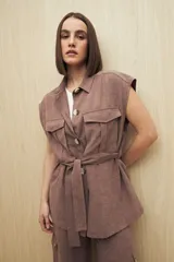 Chaleco color marrón con cuello camisero, sin mangas, cierre frontal con botones y cinturón de lazo. Presenta bolsillos de parche en el pecho.