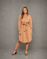 Vestido camisero color camel, de corte midi con mangas largas, cuello clásico, bolsillos frontales con solapa y cierre de botones. Incluye un cinturón de la misma tela para ajustar la cintura y presenta un ruedo redondeado con aberturas laterales.
