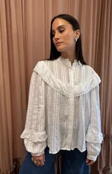 Camisa blanca de manga larga con textura a rayas verticales, canesú con volados y detalles de encaje en el pecho y el cuello alto. Cuenta con cierre frontal de botones ocultos y mangas abullonadas con volados en los puños.