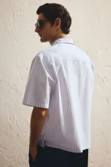 Camisa de manga corta en popelina de algodón a rayas verticales celestes y blancas. Modelo boxy con cuello inglés, tapeta clásica, bolsillo superior abierto y canesú en la espalda. Bajo recto con aberturas laterales.