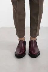 Botín asimétrico de estilo Chelsea color bordó, confeccionado en cuero vacuno, con paneles elásticos laterales negros, puntera cuadrada, suela de plataforma y tacón de bloque cuadrado.