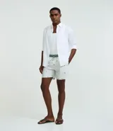Bermuda de baño para hombre confeccionada en microfibra ligera, color blanco, con una estampa sutil de cocoteros en tonos verdes/azulados. Presenta cintura elástica con cordón de ajuste contrastante en color verde, y bolsillos laterales.