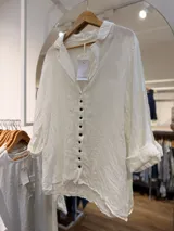 Camisa de lino color blanco, con cuello abierto, manga larga con puños vuelto y cierre frontal de botones oscuros.