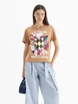 Remera de algodón color marrón con estampa frontal que incluye texto, flores, flechas y un patrón de rombos. Presenta cuello redondo y mangas cortas.
