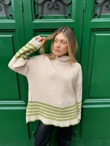Sweater beige de tejido grueso con cuello alto, corte oversize y tajo a los lados. Tiene rayas horizontales verdes en el ruedo y en los puños.
