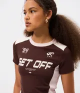 Remera de algodón con corte ajustado, manga corta y cuello redondo. Presenta un diseño bicolor en marrón y crema con estampado gráfico estilo college y lettering frontal.