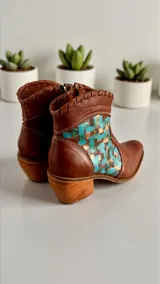 Botas de cuero color marrón tierra con taco medio, detalles de costuras artesanales en el borde superior y un aplique lateral con diseño geométrico pintado en tonos turquesa y cobrizo.