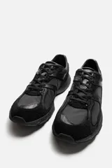 Zapatilla deportiva tipo running de color negro, perteneciente a la colección especial Origins. Presenta una parte superior confeccionada en una combinación de cuero con acabado de gamuza y tejido transpirable. Cuenta con una suela de volumen con diseño irregular y cierre mediante cordones.