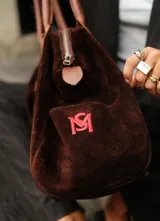 Bolso tipo duffle color verde militar, confeccionado en gamuza, con manijas de cuero marrón. Incluye bolsillos laterales, uno con clip, cierre posterior y un bolsillo interno amplio. Detalle de bandera y logo bordado en los laterales.