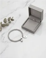 Pulsera elástica de acero compuesta por pequeñas esferas metálicas. Presenta dos dijes colgantes: uno en forma de corazón y otro rectangular con la inscripción 'Good Luck'.