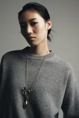 Collar largo con cadena de eslabones finos en tono dorado envejecido. El colgante consiste en un jarrón alargado dorado envejecido del cual emergen tres flores oscuras con detalles en relieve y hojas pequeñas.