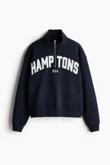 Buzo oversized azul marino con interior cepillado suave, hombros caídos, cuello elevado con medio cierre y puños y bajo acanalados. Presenta un estampado frontal con la inscripción "HAMPTONS USA" en color blanco.