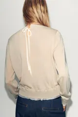 Jersey de punto color beige claro, manga larga y cuello redondo. Presenta un detalle de tejido a contraste (posiblemente una tela blanca con estampado sutil) asomando en el ruedo y el cuello.