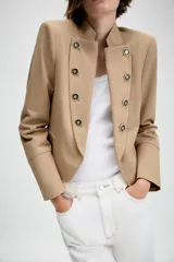 Blazer entallado de sarga color beige con cuello subido y escote en pico. Presenta manga larga con hombreras, detalle de botones metálicos decorativos en el frente y cierre frontal con gancho metálico. Incluye falsos bolsillos de vivo y abertura en la espalda.