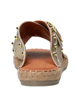Sandalias tipo ojota con tiras cruzadas en cuero metalizado dorado, decoradas con tachas metálicas y hebilla lateral. Presentan una base con detalle de yute trenzado alrededor del contorno.