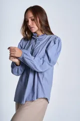 Camisa de color azul con cuello alto decorado con volados y lazo de ajuste. Presenta tablas decorativas en los hombros, cierre frontal con botones de nácar y mangas largas con puños elásticos que generan un efecto abullonado.