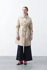 Trench de diseño clásico en color beige, con cierre frontal de botones, cinturón ajustable con hebilla a tono y mangas con trabillas en los puños.