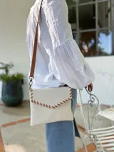 Cartera tipo sobre color crudo, confeccionada en cuero, con correa larga de cuero marrón y costuras decorativas en contraste.