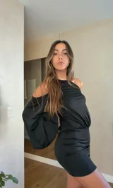 Vestido corto negro con cuello halter, hombros descubiertos y mangas largas acampanadas. Presenta un diseño ajustado en la cintura y falda con frunces laterales.
