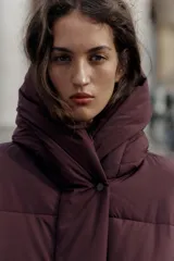 Campera puffer acolchada de color negro, con cuello envolvente, capucha integrada, manga larga, bolsillos laterales ocultos y bajo con elástico. Cierre frontal con botones a presión ocultos por solapa.