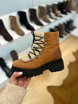 Bota tipo borcego de cuero marrón con suela track negra de goma. Presenta un aplique frontal desmontable de corderito sintético color crema y cordones negros con ganchos metálicos.