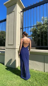 Vestido largo color marrón, con escote recto y tirantes finos. Presenta un diseño con nudo frontal que crea un efecto drapeado y una abertura central que deja ver una capa inferior más corta.