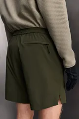 Bermuda de hombre color verde militar, de corte regular fit, confeccionada en tejido técnico compacto con elasticidad. Presenta cintura ajustable con cinturón a contraste, bolsillos frontales y un bolsillo plastrón con cierre de cremallera oculta en una de las perneras.