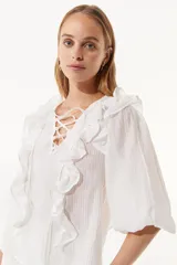 Blusa blanca de tela texturizada con volados frontales, escote en V con cordones cruzados y mangas tres cuartos abullonadas.