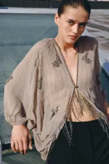 Top de viscosa color beige con escote en V, mangas murciélago abullonadas por debajo del codo y bajo asimétrico. Presenta delicados bordados con aplicaciones de abalorios y cuentas brillantes en el frente y las mangas.