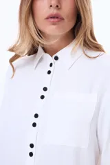 Camisa blanca de manga larga con cuello clásico y cierre frontal de botones negros en contraste. Presenta un bolsillo frontal en el pecho y un diseño de líneas simples y corte recto.