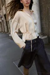 Camisa de manga larga color beige claro, con cuello solapa, escote pico y detalle de cinturón fino a tono que se anuda en la cintura.