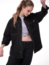 Camisa oversize de denim negro con costuras en contraste color blanco, bolsillos dobles en el pecho con solapa, cierre frontal de botones metálicos y dobladillo redondeado.