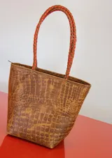 Cartera tipo tote de gamuza color marrón, con correas trenzadas y detalle de piedras naturales incrustadas en líneas verticales. Cuenta con cierre metálico y forro interior de tela.