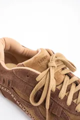 Zapatilla de perfil bajo confeccionada en gamuza combinada en tonos marrones. Presenta detalles de flecos en el lateral, costuras decorativas en forma de X a lo largo de la base y cordones al tono.