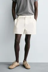 Shorts de algodón color blanco roto, de corte regular fit, con cintura ajustable mediante cinturón del mismo tejido y hebilla metálica. Presenta bolsillos delanteros y bolsillos traseros de vivo.
