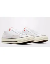 Championes Converse modelo Chuck 70 Ox, de corte bajo, confeccionados en lona blanca con detalles en gamuza gris claro en los laterales. Presentan suela de goma con franjas negras y ojal superior en color rojo.