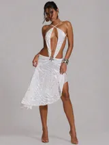 Vestido midi asimétrico blanco con escote halter cruzado, recortes profundos y espalda descubierta. Cuenta con falda de caída irregular, frunce en la cintura y aplique metálico central.