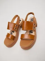 Sandalias de cuero color marrón con plataforma y taco cuadrado. Presentan dos tiras anchas sobre el empeine y una tira trasera con elástico. Decoración de doble argolla en los laterales.