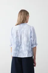 Camisa de algodón con fondo azul claro a rayas finas y estampa floral blanca en relieve. Presenta cuello cerrado, cartera de botones frontal y mangas cortas con volumen y puño elastizado. Posee frunces en el frente que aportan caída.