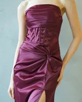 Vestido largo strapless de satén color bordó, con cuerpo estilo corsé estructurado y detalle de drapeado lateral que desemboca en una abertura frontal en la falda.